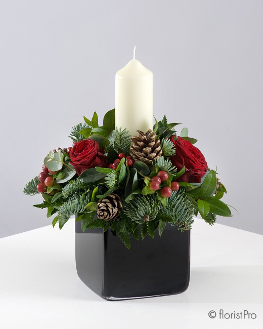 Christmas candle arrangement with red roses, fresh festive greenery, pine cones, red berries, with a pillar candle in a container handcrafted by Blooming Occasions Romford 